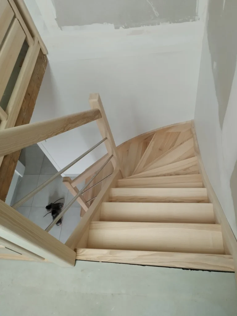 Vue plongeante depuis le palier sur un escalier quart tournant en frêne olivier, marches balancées sans contremarche, rampe Régate lisses inox — HALLOU ESCALIERS, Erbrée (35).